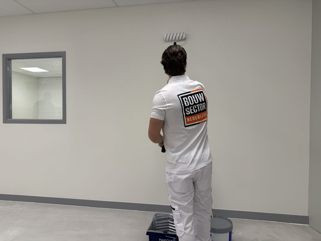 Schilder met het Bouwsector-logo op zijn rug zet de roller op een lichte wand naast een binnenraam