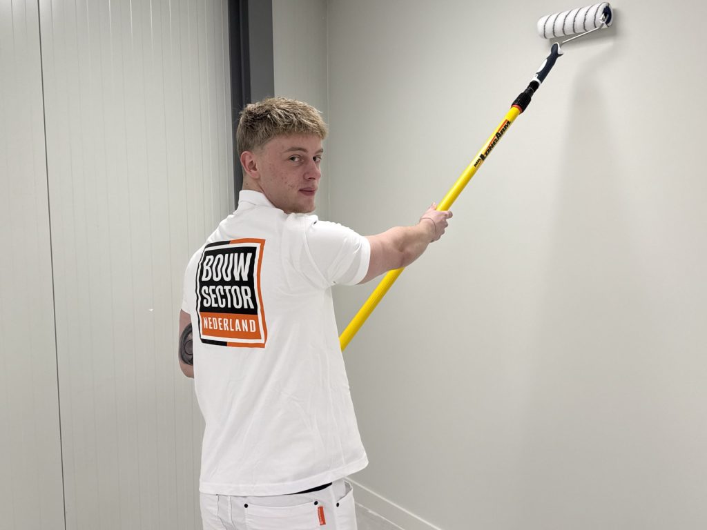 Schilder van Bouwsector Nederland kijkt om terwijl hij met een lange roller een witte wand saust