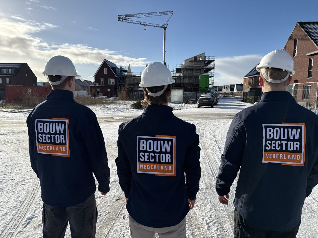 Vakmensen in donkerblauwe jassen met oranje logo voor een bouwplaats onder een blauwe lucht