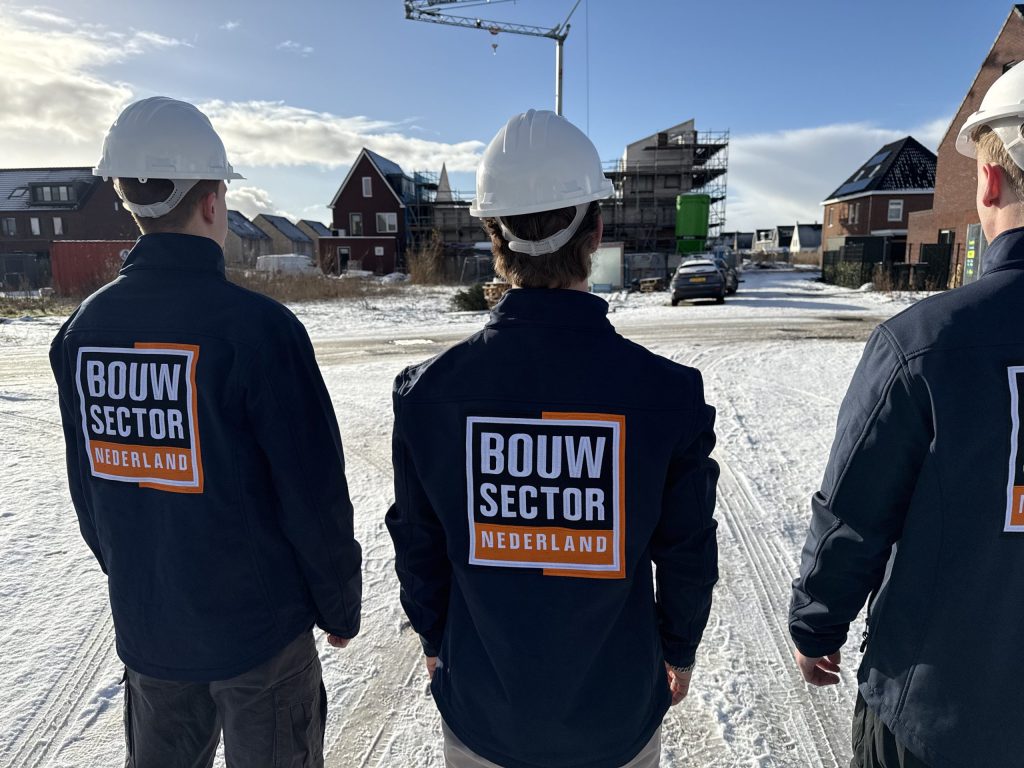 Drie vakmensen van Bouwsector Nederland met het logo op de rug voor een rij nieuwe huizen